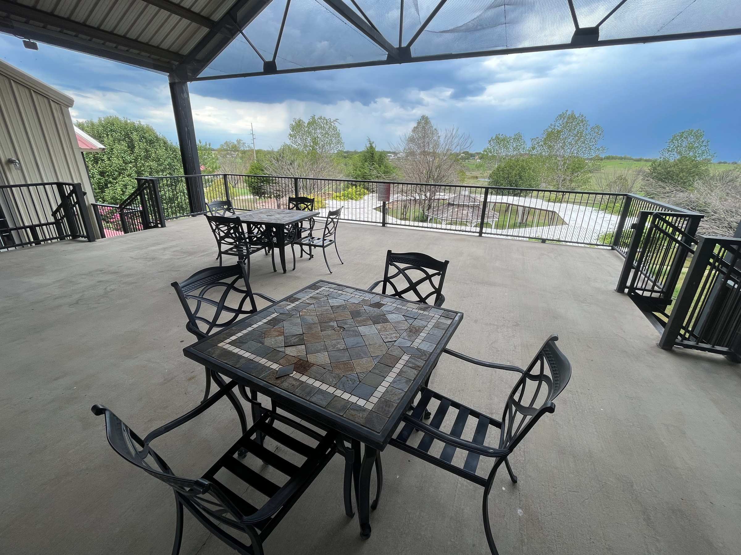 The covered upstairs patio: a slate-topped table and iron chairs, with a rail looking out over trees and the grounds under a stormy sky.