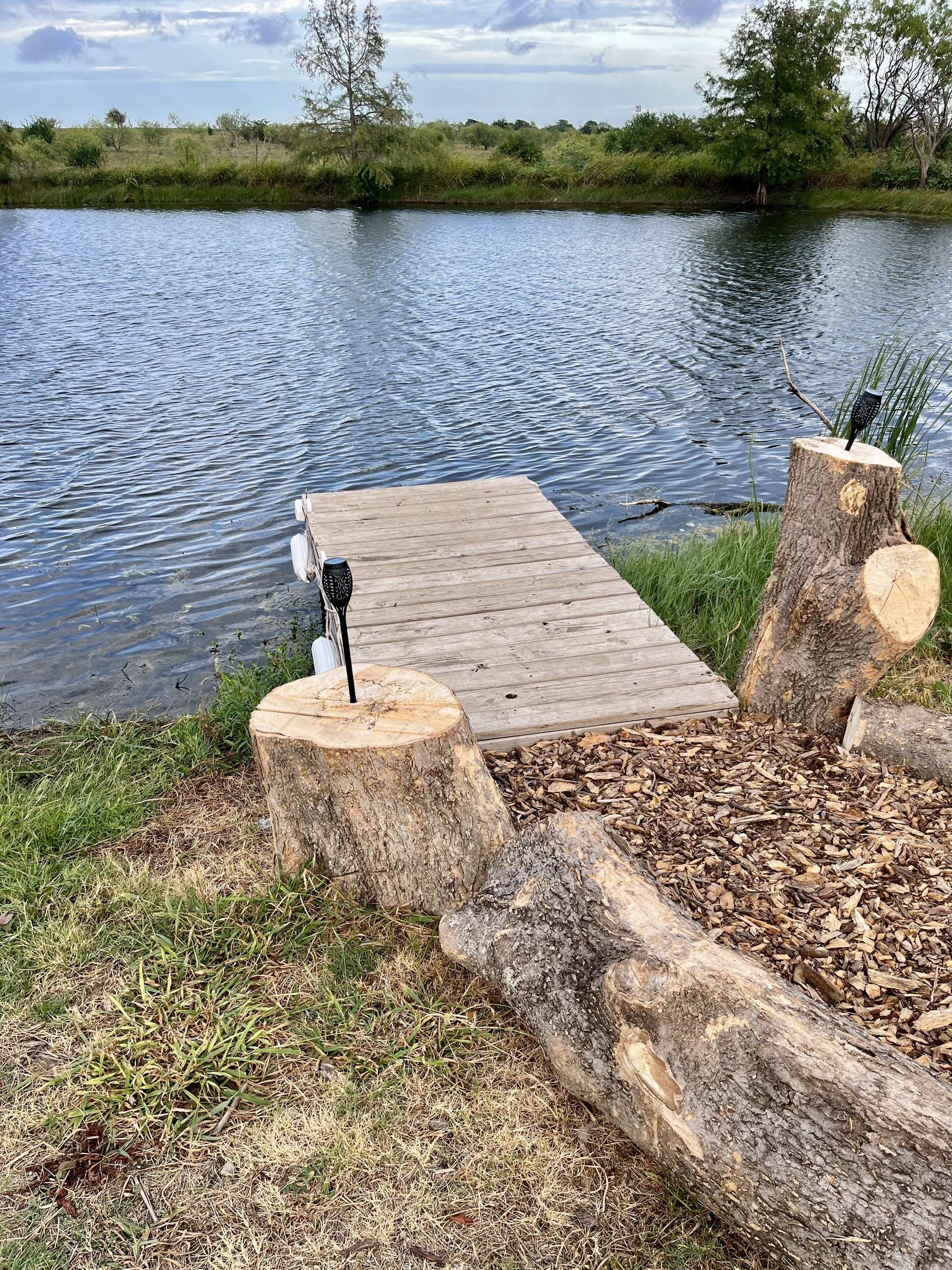 A small wooden fishing dock on the lake, with tree-stump seats and a log along a wood-chip path to the water.
